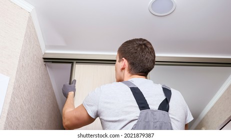 Employee furniture assembler in white t-shirt and jumpsuit checks wooden sliding door of modern wardrobe with mirror close back low angle shot