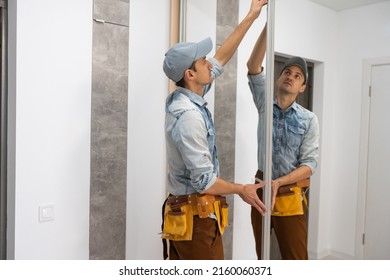 Employee furniture assembler in white t-shirt and jumpsuit checks wooden sliding door of modern wardrobe with mirror.