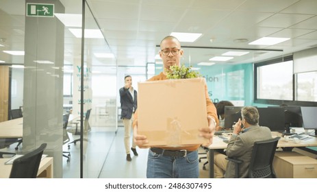 Employee with glasses carrying a box of belongings, walking through a modern office after being dismissed from work, with colleagues in the background. - Powered by Shutterstock