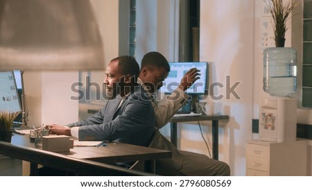 Employee having fun at work during nightshift, bumping into coworker using desk chair, with friends racing eachother to avoid boredom during overnight stay in office.
