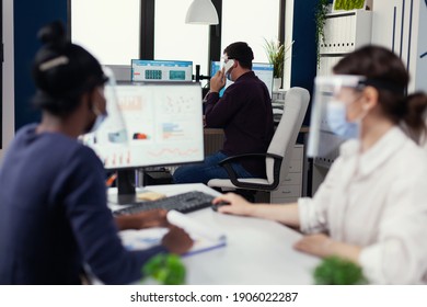 Employee having a phone conversatiion with client sitting at desk wearing face mask for covid19. Multiethnic team working in company with new normal respecting social distance during global pandemic - Shutterstock ID 1906022287