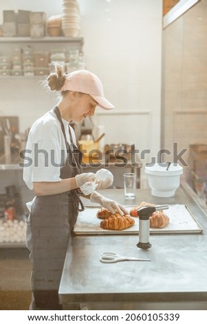 Employee holds confectioner sleeve with delicious cream to decorate croissants in bakery shop