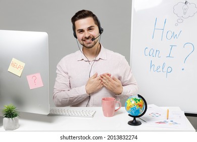 Employee kind operator business man in set microphone headset for helpline assistance sit work at call center office desk with pc computer put folded hands on heart isolated on grey background studio