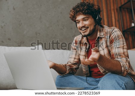 Employee operator business Indian man in set microphone headset fot helpline assistance sit work laptop pc computer at call center on grey sofa couch stay at home hotel flat spend time in living room