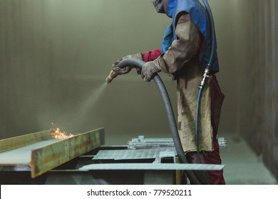 An employee prepares a metal part for painting. A harsh man works in the factory. Sandblast. Blast it.