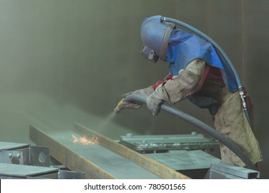 An employee prepares a metal part for painting. A harsh man works in the factory. Sandblast. Blast it.