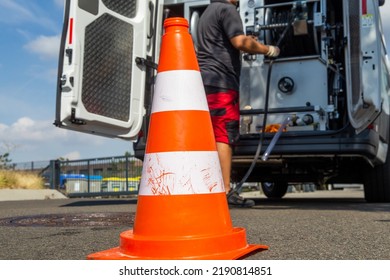 Employee Of A Sewer Cleaning Company Cleaning A Street Drain. In The Foreground, The Construction Site Cap, In The Background, The Emergency Vehicle