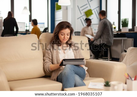 Employee taking break sitting on confortable couch holding tablet browsing, reading great news and smiling. Multiethnic creative coworkers planning new financial project working in open plan office
