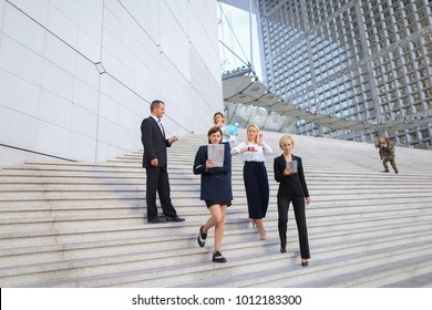 Employees Of Large  Corporation Walking In   And Speaking By Smartphone. Concept Of Successful Business Partners Outside. Young Prosperous People Dressed In Black Suits Look Happy.