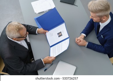 Employer interviewing a young male job seeker for a business vacancy sitting at a table reading his CV viewed from above