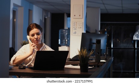 Empowered Female Manager Talking On Landline Phone Call, Working Late At Night To Plan Report Presentation On Laptop. Using Financial Data Research To Work On Executive Shares.