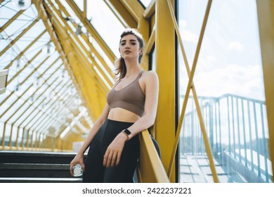 Empowered young Caucasian woman with water bottle, leaning on railing in a bright yellow urban pedestrian bridge, reflecting wellness and a confident active lifestyle - Powered by Shutterstock