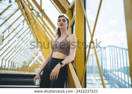 Empowered young Caucasian woman with water bottle, leaning on railing in a bright yellow urban pedestrian bridge, reflecting wellness and a confident active lifestyle