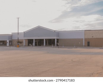 An empty abandoned big box storefront building with a blank gabled sign space and a vacant asphalt parking lot under a hazy sky