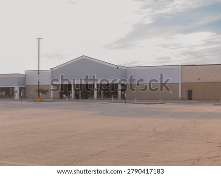 An empty abandoned big box storefront building with a blank gabled sign space and a vacant asphalt parking lot under a hazy sky