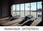 Empty airport terminal seating area with a large window view of the runway and an airplane taking off in the background. Bright sunlight casting shadows on the carpeted floor