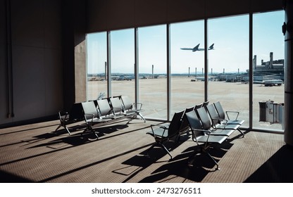 Empty airport terminal seating area with a large window view of the runway and an airplane taking off in the background. Bright sunlight casting shadows on the carpeted floor - Powered by Shutterstock