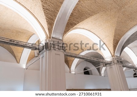 An empty, arched ceiling room in an old European city, illuminated by filtered natural light The space features a horizontal beam and textured brickstone ceiling contrasting with white walls Docum