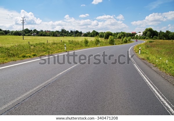 Empty asphalt road with a double bend between\
flower meadows in the countryside. Red car on the road in the\
distance. Village among the green leafy trees background. Sunny day\
with blue skies.