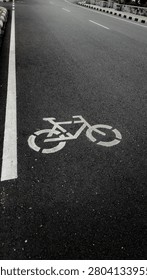 An empty asphalt road features a white bicycle lane symbol painted on dark pavement. Clear white road markings and curbside borders indicate a designated, quiet cycling path under calm daylight conditions.