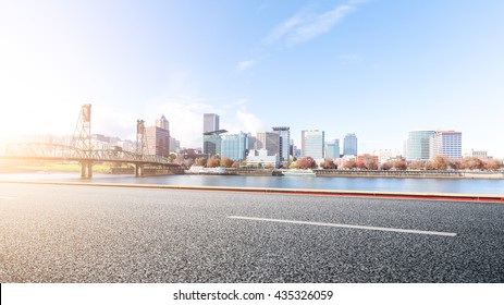 Empty Asphalt Road Near Water With Cityscape And Skyline Of Portland At Sunrise