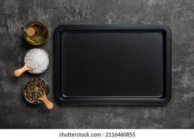 Empty baking sheet, olive oil jar, salt and pepper grains over black textured background. Rectangular oven tray and spices for baking and roasting. Baking pan for cooking and food design. Copy space.