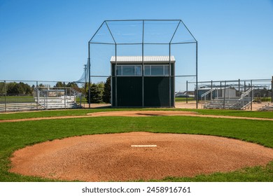 Empty baseball diamond with backstop and pitcher's mound. Blue sky and green grass. Small town baseball field. Red clay soil with green grass and bleachers. Announcer's booth in background,  - Powered by Shutterstock