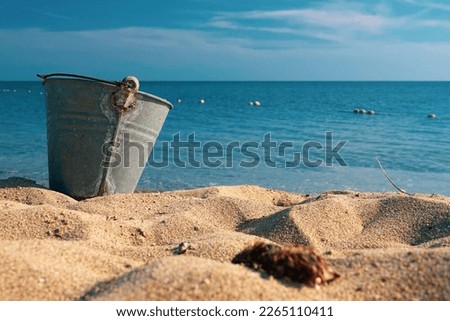 An empty beach metal bucket or bin on the beach among sands with blue sea and sky as background. 1980's summer concept. Water pollution