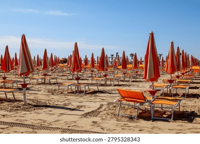 Empty beach with rows of closed orange and white umbrellas and lounge chairs on a sunny day, the sandy shore shows tire tracks and footprints, recent activity or preparation for visitors