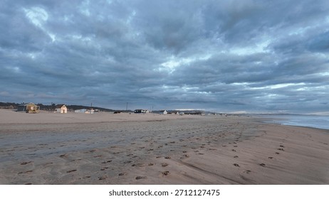 An empty beach under moody clouds, footprints marking the untouched morning sand.
