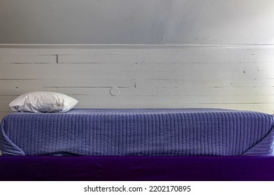 An Empty Bed Covered In Purple Blanket With White Pillow In Front Of Rustic Wooden Wall.