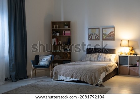Empty bedroom with electric lamp on shelves, cozy armchair, pictures above bed at night