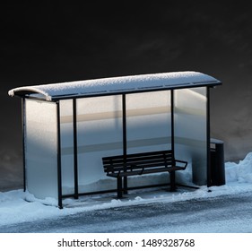 Empty bench in bus stop in winter, with dark sky in background and some dim light from low sun.