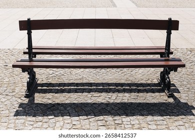 Empty bench on cobblestone square. Classic wooden seat with iron legs. Bright daylight and sharp shadow. Urban public park element. Solitary bench in open space. Peaceful outdoor seating.