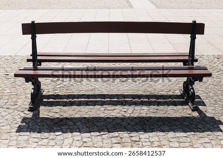Empty bench on cobblestone square. Classic wooden seat with iron legs. Bright daylight and sharp shadow. Urban public park element. Solitary bench in open space. Peaceful outdoor seating.