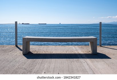 Empty bench on concrete deck, sea view. Cargo ships moored, blue sky background. Industrial harbor of Drapetsona, Greece - Powered by Shutterstock