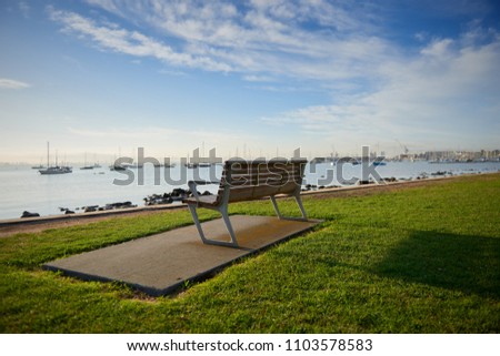 An empty bench on grass by the waters edge in the inspiring bright morning sun with a gorgeous view of boats and marina in the distance.