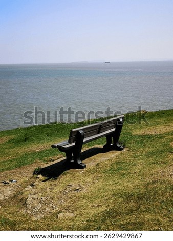 Empty bench on a grassy cliff, Friars Point, overlooking Whitmore Bay, on a cloudless, blue sky day, with a distant ship and island just visible. Barry, Vale of Glamorgan, Wales, United Kingdom 