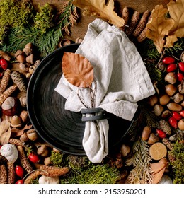 Empty black ceramic plate with cloth napkin over ambiance magic autumnal forest background. Autumn leaves, moss, fir cones, snail shell over wooden surface. Thanksgiving menu. Flat lay. Square image