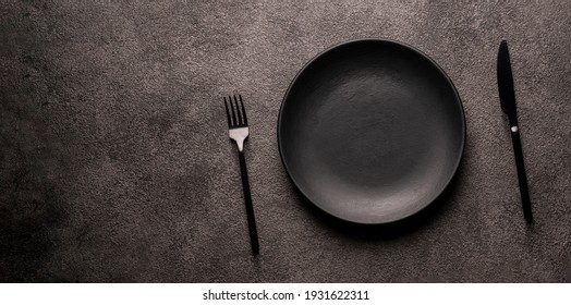 An Empty Black Plate And A Fork With A Knife On A Dark Background. Flatlay Photo, Mockup Concept For Advertising And Design Of Fast Food Restaurants, Copy Space