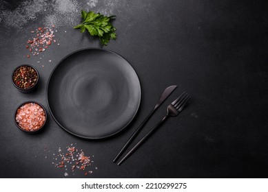 An empty black plate on a dark concrete background with spices, herbs and cutlery. Home dinner cooking - Powered by Shutterstock