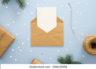 Empty Blank Card In Kraft Paper Envelope On Pastel Blue Background Decorated With Confetti Star, Gift Box, Wrapping Paper And Fir Tree Branches. Christmas, New Year, Winter Holiday Invitation Mockup.