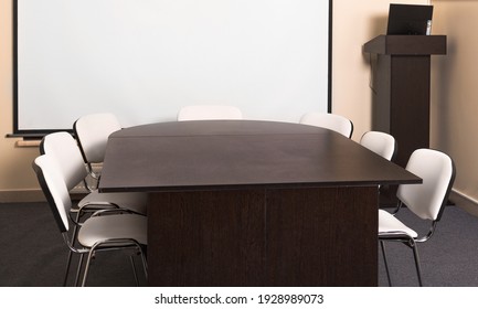 Empty Board Room. Small Conference Room With Table And Chairs. Wall Projector Screen And Lectern With Laptop. A Room Without People. No Windows