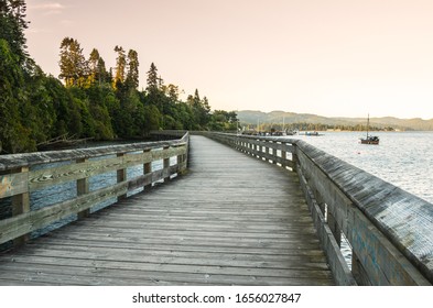 Empty boardwalk along the the shore of a bay at sunset. Some jetties with moored boats are visible in background.
