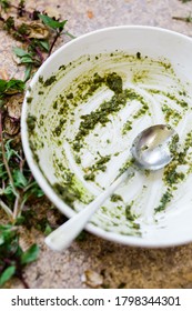 An Empty Bowl With Leftover Basil Pesto Sauce And A Dirty Spoon. Vertical Photo. In The Background, You Can See Basil Leaves And Stem On A Dirty Countertop.