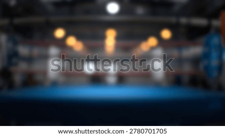 An empty boxing ring with a blue mat and ropes is illuminated by overhead lights in a sports arena.