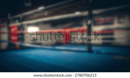 An empty boxing ring with a blue mat and red ropes is shown in a dimly lit gym interior.