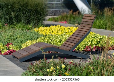 Empty brown wooden deck chair or chaise longue on tile among decorative grass and flowers in recreation area. Garden landscape with chairs in city park. Concept of recreation, tanning in yard
