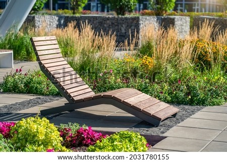 Empty brown wooden deck chair or chaise longue on tile among decorative grass and flowers in recreation area. Garden landscape with chairs in city park. Concept of recreation, tanning in yard
