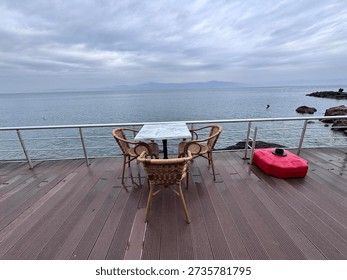 Empty cafe table and chairs on a wooden terrace overlooking a calm sea under cloudy sky. Peaceful coastal lifestyle scene with wide copy space, ideal for travel, relaxation, and seaside concepts.